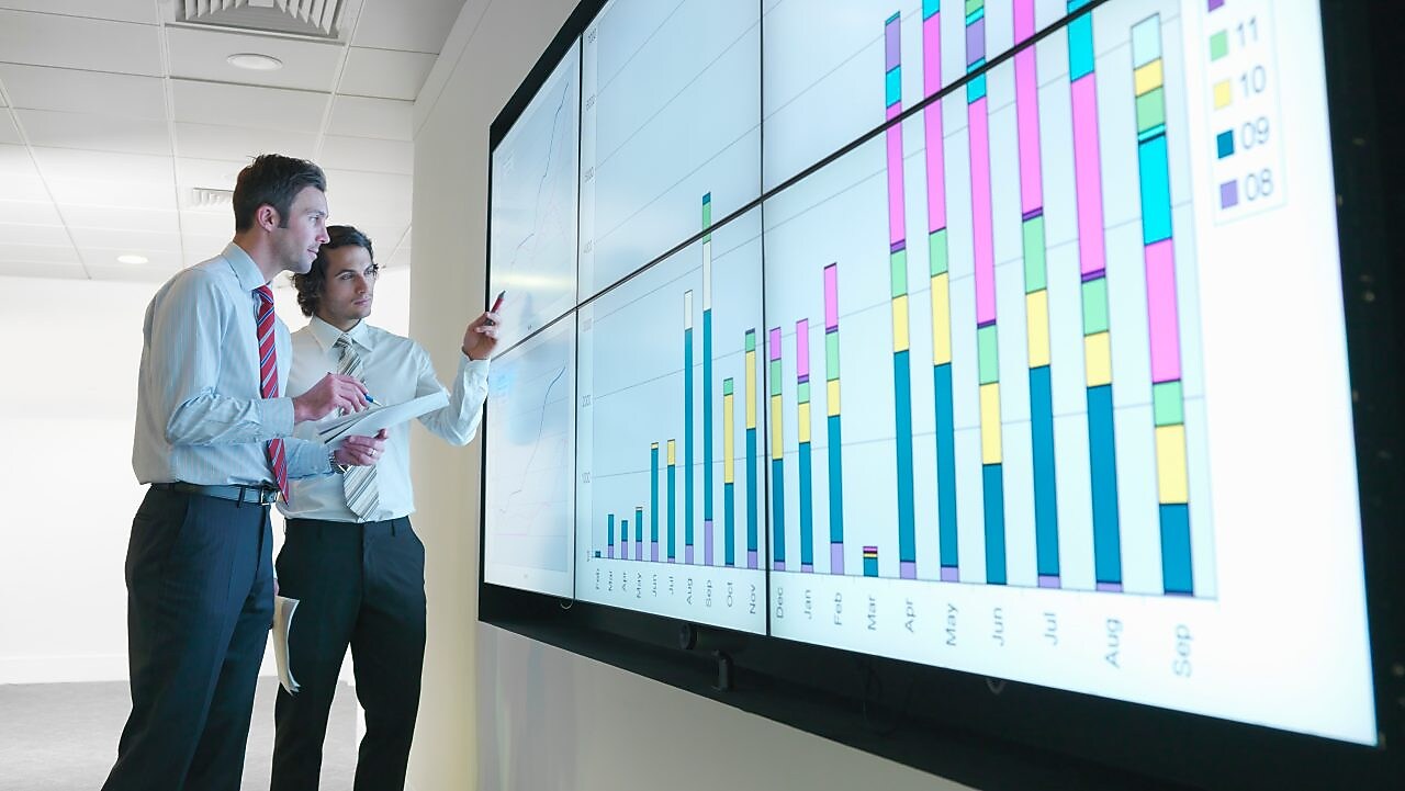 Business men analyse reports displayed on a large digital screen